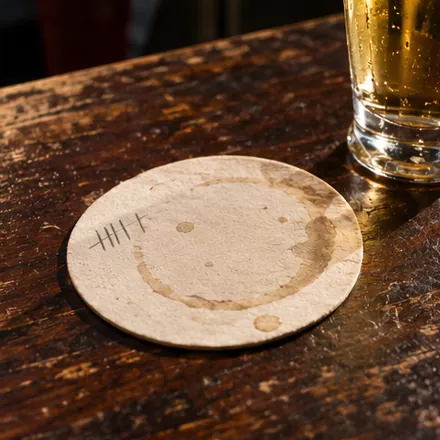 A worn Kölsch coaster on a dark pub table, five pencil marks on its rim